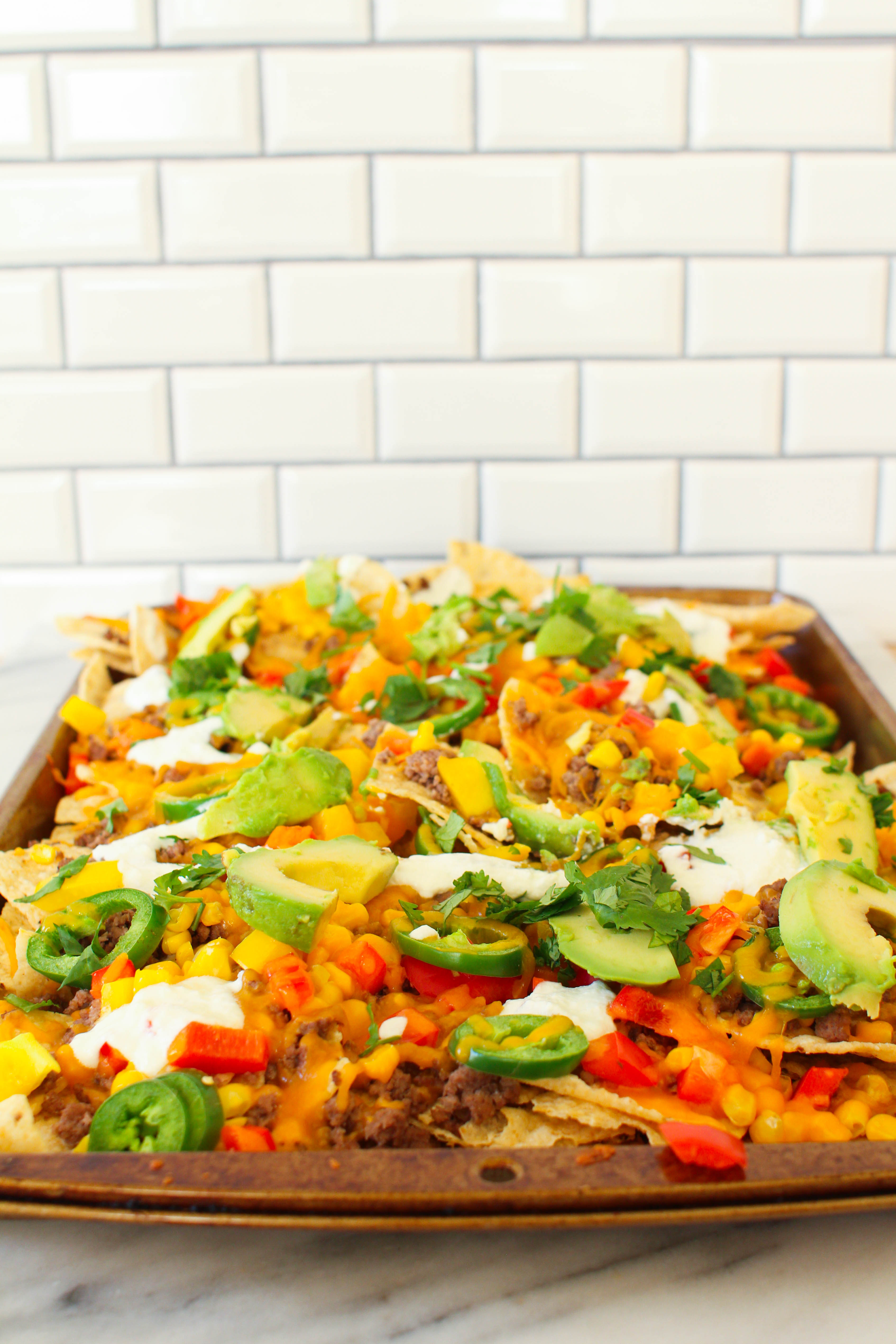 Perfect for game day, these cheesy beef sheet pan nachos can feed a crowd. Tortilla chips topped with seasoned ground beef, corn, peppers, jalapenos and cheese and baked until crispy and melted. Topped with sour cream, avocado and cilantro. 